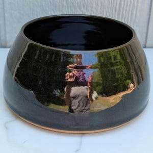 May include: A glossy, dark brown ceramic bowl with a wide, rounded shape. The interior is a deep black, and the exterior reflects the surrounding environment. The bowl sits on a light brown base, showcasing a handcrafted aesthetic.