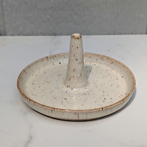 May include: A circular ceramic ring holder featuring a central conical post. The piece has a speckled off-white glaze with brown flecks throughout and a smooth, shallow dish base with a defined rim, showcasing a natural, earthy aesthetic.