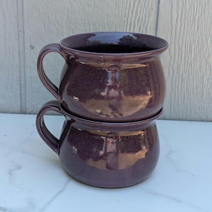 May include: Two stacked, deep purple ceramic mugs with rounded bodies and curved handles. The mugs have a speckled texture and a glossy finish. The mugs are designed for beverages and are suitable for kitchen use.