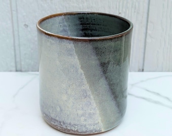 May include: A cylindrical ceramic tumbler featuring a reactive glaze with a blend of muted blue, grey, and white tones. The rim is defined by a thin, dark brown band, and the exterior surface displays a smooth, mottled texture with color variations.