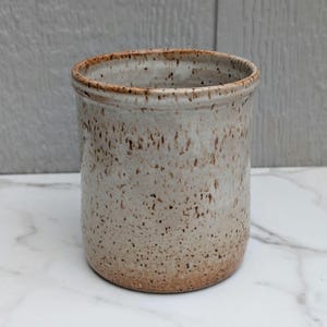 May include: A cylindrical ceramic utensil holder featuring a speckled off-white glaze with brown flecks throughout. The rim and base exhibit a subtle gradient of warm, earthy brown tones, creating a textured appearance on the exterior surface.