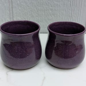 May include: Two handmade, speckled purple ceramic cups. The cups have a rounded shape with a slightly flared rim. The exterior is a deep purple with lighter purple speckles, and the interior is a darker shade of purple.
