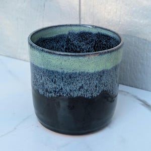 May include: A cylindrical ceramic cup featuring a two-tone glaze. The upper portion is a light, speckled mint green, which transitions into a deep, solid black base through a textured, mottled gradient of dark blue and grey tones.