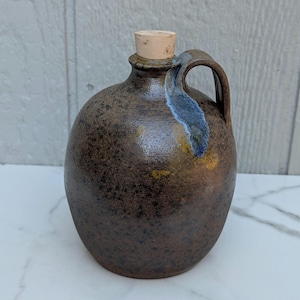 May include: A round ceramic jug with a dark brown speckled glaze and a single side handle. The neck features a blue-toned glaze accent near the handle attachment. The jug is topped with a light brown cylindrical cork stopper.