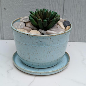 May include: A light blue ceramic planter with a matching saucer. The planter is filled with smooth, light-colored stones and a dark green succulent. The planter has a speckled texture and a slightly flared rim.