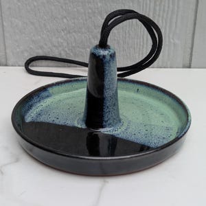 May include: A handmade ceramic incense holder with a black and teal glaze. The holder features a central cone with a black cord loop and a circular base. The base is divided into black and teal sections.