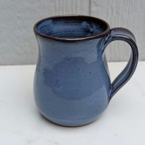 May include: A handmade blue ceramic mug with a dark brown rim and handle. The mug has a rounded body and a curved handle, perfect for enjoying your favorite beverage. The mug is approximately 4 inches tall.