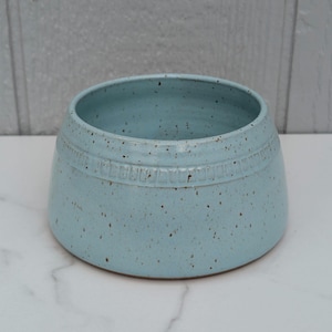 May include: A light blue ceramic bowl with a speckled finish. The bowl has a wide, rounded shape with a decorative band around the middle. The interior is a lighter shade of blue.