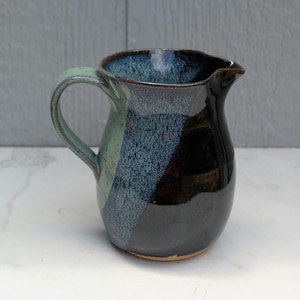 May include: A ceramic pitcher featuring a glossy finish with a color palette of deep black, mottled blue, and light sage green. The vessel has a curved handle on the left side, a wide opening with a small pouring spout, and a rounded body shape.