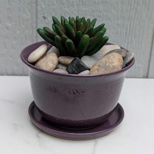 May include: A purple ceramic planter with a matching saucer, filled with smooth stones and a dark green succulent plant. The planter has a rounded shape and a textured surface. The stones are various colors, including white, gray, and black.