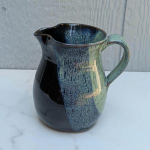 May include: A ceramic pitcher featuring a two-tone glaze with a solid glossy black section on one side and a speckled blue and light green finish on the other. The vessel has a curved handle, a rounded body, and a distinct pouring spout at the top.