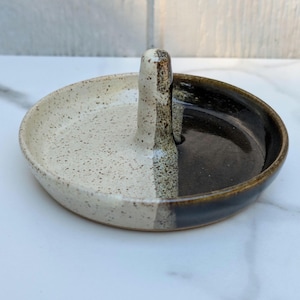 May include: A circular ceramic ring dish featuring a central vertical post. The dish is divided into two distinct sections: one side is a speckled off-white glaze, while the other side is a smooth, glossy dark brown glaze, meeting at the center post.