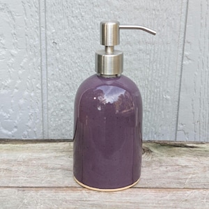 May include: A purple ceramic soap dispenser with a stainless steel pump. The dispenser has a rounded shape and a light beige base. The background is a weathered wooden surface and a gray wooden wall.