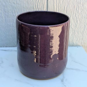 May include: A cylindrical ceramic cup with a deep purple, glossy glaze and subtle speckled texture. The vessel features a smooth, rounded body and a wide, open top, showcasing a reflective surface that catches light across its dark, uniform exterior.