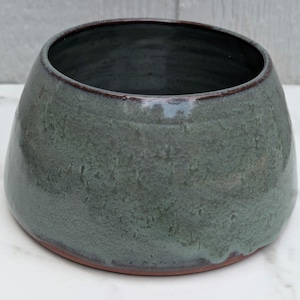 May include: A wide, round ceramic bowl with a tapered silhouette and a thick, dark rim. The exterior features a mottled, textured glaze in shades of sage green and slate gray, while the base reveals a contrasting reddish-brown clay material.