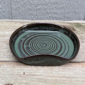 May include: A handmade ceramic spoon rest. The oval-shaped rest features a dark brown rim and a teal-green interior with a spiral design. The spoon rest is placed on a wooden surface.