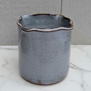 May include: A cylindrical ceramic vessel with a light blue-grey speckled glaze and a scalloped rim. The top edge of the container features a thin, dark brown accent line that contrasts with the smooth, muted blue body of the pottery piece.