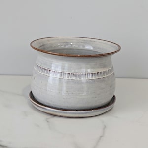 May include: A handcrafted, light gray ceramic planter with a matching saucer. The planter features a decorative band of white rectangular patterns around the middle and a brown rim. The design is simple and elegant, suitable for indoor plants.