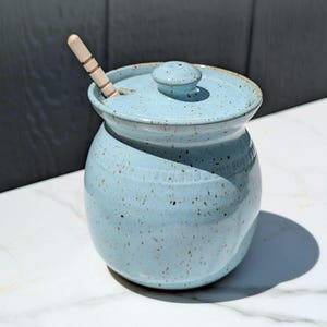 May include: A light blue ceramic jar with a lid and a wooden spoon. The jar has a round shape with a speckled pattern. The lid has a small knob on top. The wooden spoon is partially inserted into the jar.