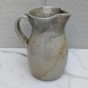 May include: A ceramic pitcher with a speckled gray and beige glaze. The vessel features a rounded body, a curved handle on the left side, and a distinct pouring spout at the top rim. The surface texture appears mottled with subtle brown accents.