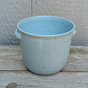 May include: A light blue ceramic planter with two small handles. The speckled surface has brown flecks. The interior is a lighter shade of blue. The planter is sitting on a wooden surface.