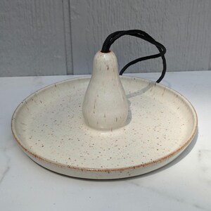 May include: A round, off-white ceramic ring dish featuring a speckled brown pattern and a central, pear-shaped upright post. A black braided cord extends from the top of the central post, providing a functional element to this ceramic accessory.