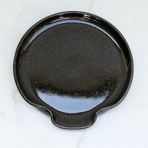May include: A black ceramic spoon rest with a glossy finish. The round rest has a small, curved indentation for holding a spoon or other utensil. The surface has a speckled texture.