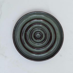 May include: A circular ceramic soap dish featuring a series of concentric raised rings. The surface has a mottled sage green glaze with dark brown accents along the ridges and the outer rim, creating a textured appearance for the round dish.