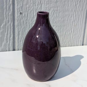 May include: A glossy, deep purple ceramic vase with a rounded body and a narrow neck. The vase has a smooth, reflective surface, and casts a shadow on the white surface it rests on. The vase is a decorative item.