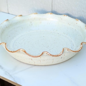 May include: Off-white ceramic pie dish with a speckled finish and a wavy, scalloped rim. The rim has a brown, rustic edge. The dish is round and appears to be handmade, suitable for baking and serving pies.