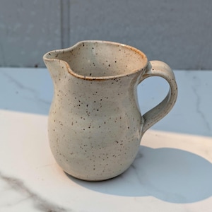 May include: A speckled, off-white ceramic pitcher with a curved handle and a pouring spout. The pitcher has a rounded body and a slightly textured surface. The rim and handle have a light brown color. The pitcher is suitable for serving beverages.