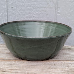 May include: A handmade, dark teal ceramic bowl with a wide, rounded shape. The bowl has a slightly textured surface and a dark brown rim. The bowl is likely used for serving food or as a decorative piece.