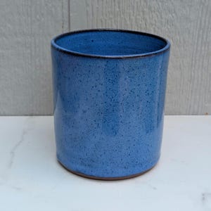May include: A cylindrical ceramic tumbler featuring a speckled blue glaze with a dark brown rim and base. The vessel has a smooth, straight-sided profile and a uniform finish, showcasing a deep cobalt blue color with subtle darker flecks throughout.