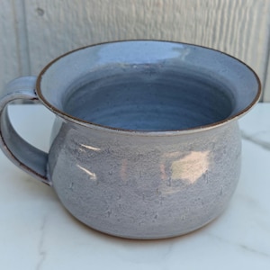 May include: A ceramic mug with a rounded, bulbous body and a wide, flared rim. The mug features a mottled, light blue-grey glaze with a smooth finish. A single curved handle is attached to the side, and the rim is accented with a thin brown edge.