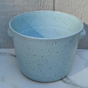May include: A light blue ceramic bowl with a speckled finish and two small side handles. The bowl has a wide, open top and a rounded base, featuring a consistent pale blue glaze with dark brown flecks distributed across the entire surface.