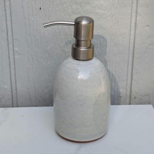 May include: A soap dispenser with a rounded, off-white ceramic body featuring subtle grey speckles and a smooth, matte finish. The top is equipped with a brushed stainless steel pump mechanism that extends outward to the left for dispensing liquid soap.