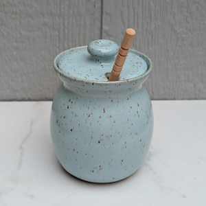 May include: A light blue ceramic honey pot with a speckled brown finish, featuring a matching lid with a small knob handle and a light-colored wooden honey dipper inserted through a circular opening in the lid, creating a functional kitchen item.