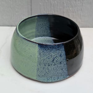 May include: A round ceramic bowl featuring a gradient glaze that transitions from a solid light sage green on one side to a speckled blue-grey in the center, ending in a deep glossy black on the opposite side with a smooth, curved rim.