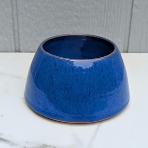 May include: A deep blue ceramic bowl with a wide, tapered base and a smooth, glossy finish. The bowl features subtle variations in color intensity across its surface, with a dark, circular opening at the top and a solid, rounded exterior shape.