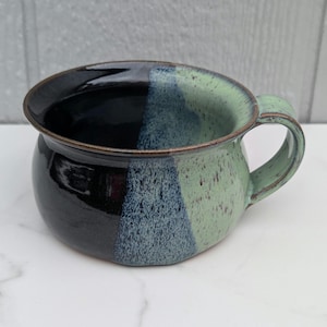 May include: A handmade ceramic mug with a unique color block design. The mug features a black, blue, and green color scheme. The mug has a rounded shape with a handle and a wide rim. The mug is suitable for coffee, tea, or soup.