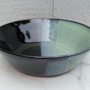 May include: A round ceramic bowl featuring a glossy finish with a distinct color-blocked design. One half of the bowl is solid black, while the other half transitions through a speckled light blue into a soft, pale green hue.