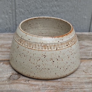May include: A speckled off-white ceramic bowl with a wide, tapered base and a narrow opening. The exterior features a decorative band of vertical indentations encircling the upper portion, with a smooth, glossy finish and dark brown speckles.