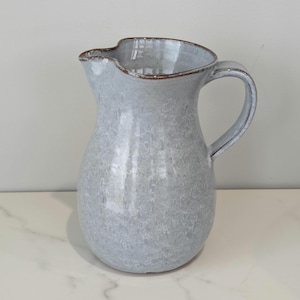 May include: A light blue ceramic pitcher with a textured surface and a curved handle. The rim and handle edges have a brown trim. The pitcher is designed with a wide opening and a slightly irregular shape, suitable for serving beverages or as a decorative piece.