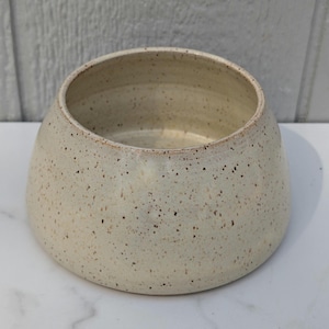 May include: A speckled, off-white ceramic bowl with a rounded shape. The bowl has a wide opening and a slightly tapered base. The exterior is covered in small, dark brown speckles, giving it a rustic appearance. The interior is a lighter shade of off-white.