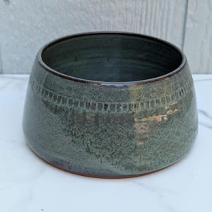 May include: A round ceramic bowl with a tapered silhouette, featuring a mottled dark green and grey glaze. The exterior surface displays a subtle horizontal band of repeating vertical dash patterns near the rim, with a smooth, dark brown base.