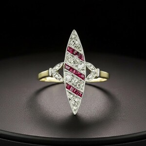 May include: An Art Deco-style ring with a central, elongated design. The ring features alternating rows of small, sparkling diamonds and vibrant red gemstones. The band is gold-toned, with decorative accents on either side of the central design.
