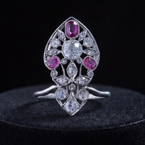Peut inclure: Une bague argentée dotée d'une monture en filigrane allongée en forme de bouclier. Le motif est centré sur une grande pierre ronde transparente, rehaussée de trois rubis roses taille ovale et de plusieurs petites pierres rondes transparentes.