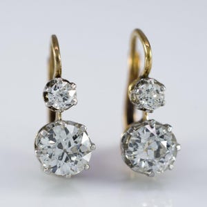 May include: A pair of gold drop earrings, each with two clear, round-cut diamonds. The diamonds are set in silver-coloured metal and hang from a gold hoop. The earrings are shown against a white background.