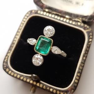 Op de afbeelding: Een vintage ring met smaragd en diamanten, gepresenteerd in een juwelendoosje. De ring heeft een rechthoekige smaragd als middensteen met een gouden rand, omringd door vier diamanten: twee rond en twee peervormig.