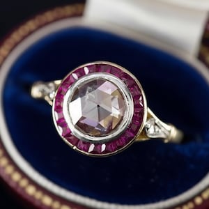 Puede incluir: Un anillo de compromiso vintage con una gran piedra central facetada rodeada por un halo de pequeños rubíes cuadrados. El anillo tiene una banda de oro y está colocado en un joyero de terciopelo azul.
